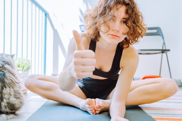 Junge sportliche Frau mit zerzausten, lockigen braunen Haaren sitzt auf einer Yogamatte und zeigt super Zeichen für Heimtraining