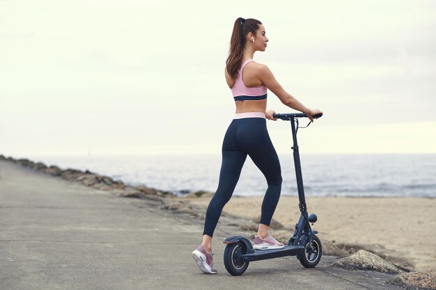 Junge sportliche Frauen in Activewear fahren an bewölkten Tagen mit ihrem Elektroroller an der Küste entlang.