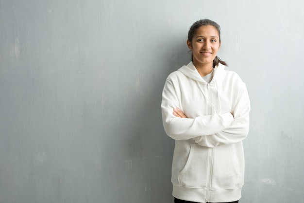 Junge sportliche indische Frau gegen eine Turnhallenwand, die seine Arme kreuzt, lächelt und glücklich