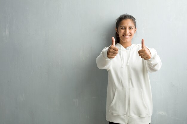 Junge sportliche indische Frau gegen eine Turnhallenwand nett und aufgeregt, sie lächelnd und anheben