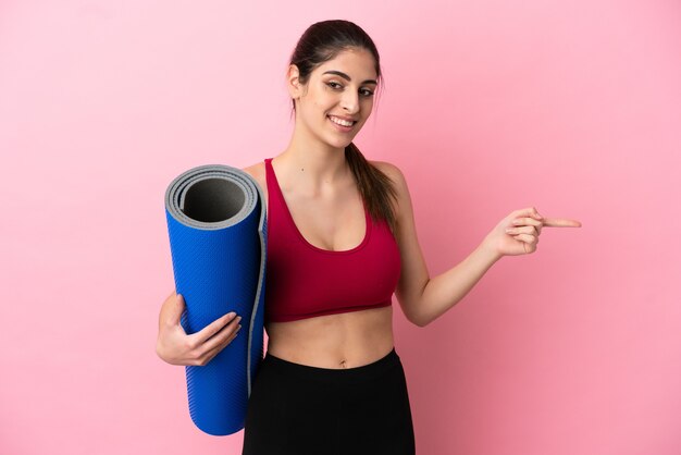 Junge sportliche kaukasische Frau, die zu Yoga-Kursen geht, während sie eine Matte hält, die mit dem Finger zur Seite zeigt