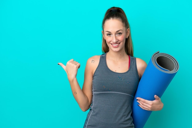 Junge sportliche kaukasische Frau, die zu Yoga-Kursen geht, während sie eine Matte isoliert auf blauem Hintergrund hält, die zur Seite zeigt, um ein Produkt zu präsentieren