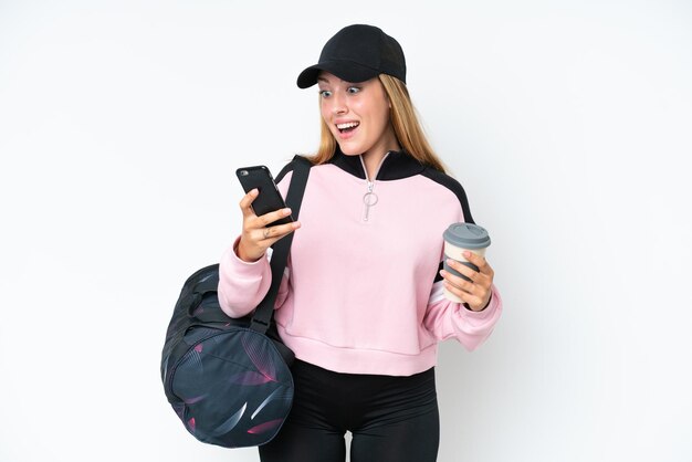 Junge sportliche kaukasische Frau mit Sporttasche isoliert auf weißem Hintergrund mit Kaffee zum Mitnehmen und einem Handy