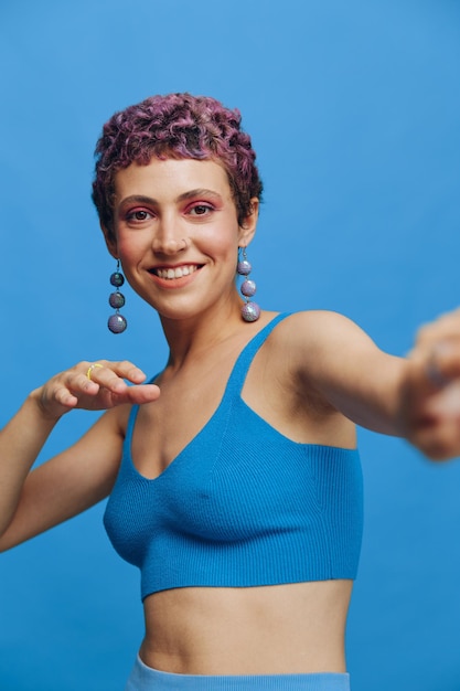 Junge sportliche Modefrau mit gefärbten Haaren und Kurzhaarschnitt posiert und tanzt in blauer Sportkleidung, lächelt und blickt auf einem blauen monochromen Hintergrund in die Kamera