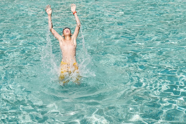 Junge springt im Schwimmbad mit erhobenen Händen aus dem Wasser, während er für ein Foto im Wasserpark posiert