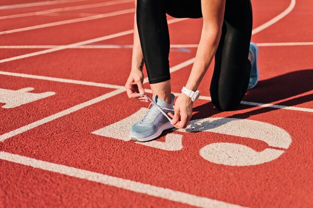 Junge Sprinterfrau in Sportbekleidung, die Schnürsenkel vor einem Rennen auf einer rot beschichteten Stadionstrecke an einem sonnigen hellen Tag bindet