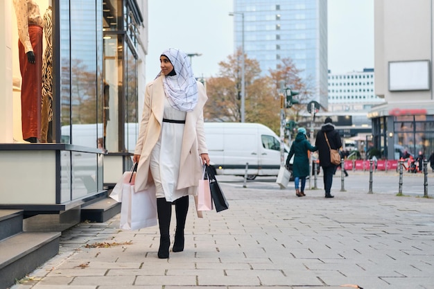 Junge stilvolle arabische Frau im Hijab, die mit Einkaufstüten durch die Stadtstraße geht