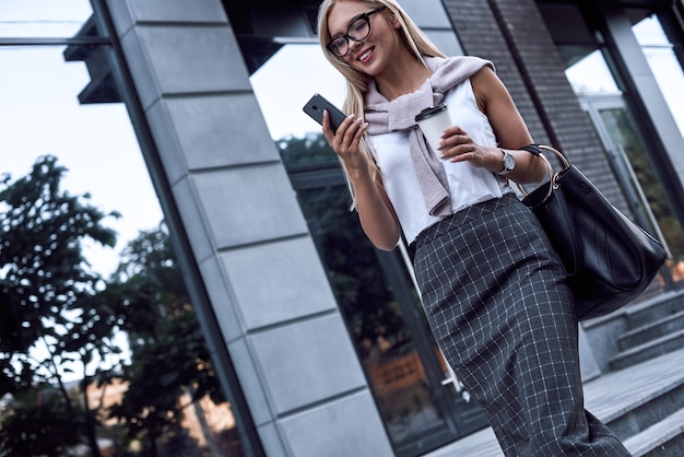 Junge stilvolle Frau, die am Smartphone plaudert, geht sie mit Morgenkaffee