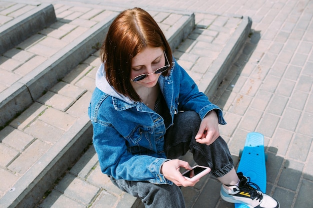 Junge stilvolle Frau geht mit einem Skateboard auf der Straße und benutzt ein Smartphone