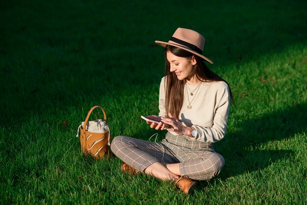 Junge stilvolle Frau im Hut benutzt Handy, während sie am Sommermorgen auf grünem Rasen sitzt.