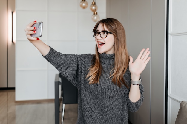 Junge stilvolle Frau in den Gläsern, die Hand winken und Hallo zum Telefonbildschirm sagen