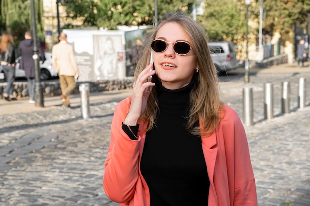 Junge stilvolle Frau in der Korallenjacke und in der Sonnenbrille, die auf einem Handy sprechen