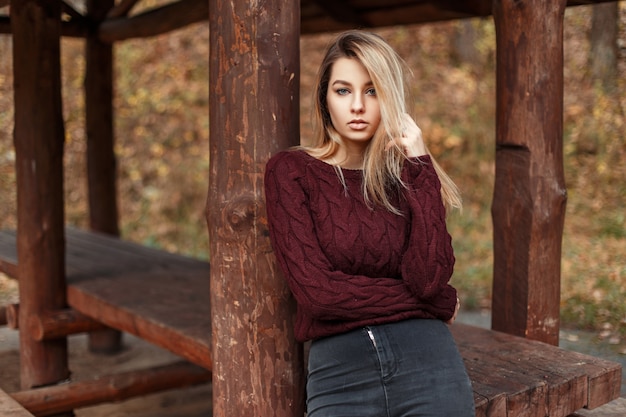 Junge stilvolle Frau in einem trendigen Weinlesepullover wirft nahe Holzsäulen am Herbsttag auf