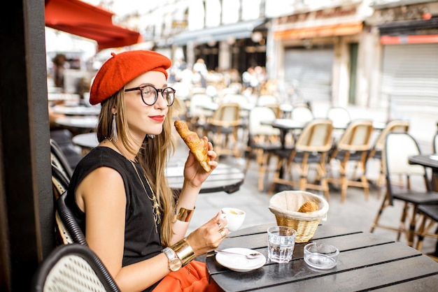 Junge stilvolle Frau in rotem Barett, die ein französisches Frühstück mit Kaffee und Croissant im Freien auf der Café-Terrasse hat?