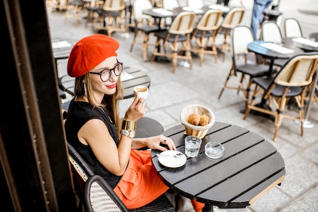 Junge stilvolle Frau in rotem Barett, die ein französisches Frühstück mit Kaffee und Croissant im Freien auf der Café-Terrasse hat?