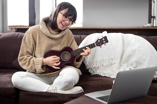 Junge stilvolle Frau mit Brille lernt, die Ukulele zu spielen. Konzept der Online-Bildung, Heimunterricht.