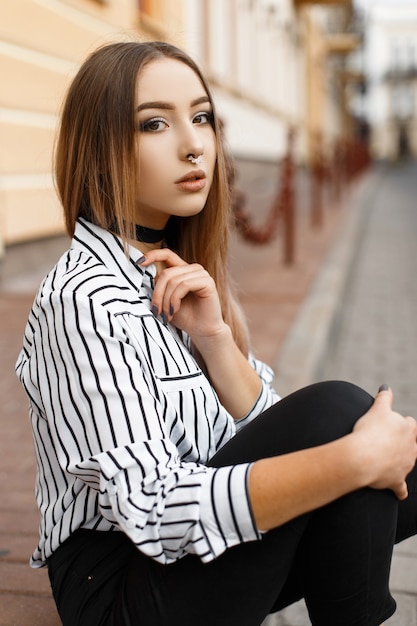 Junge stilvolle Frau mit einem Ohrring in der Nase in modischen schwarzen Jeans in einer gestreiften Bluse ruht auf dem Boden in der Stadt in der Nähe von Vintage-Gebäuden.