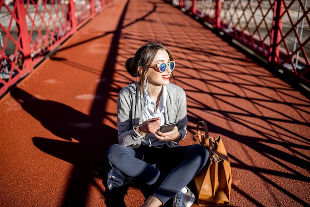 Junge stilvolle Geschäftsfrau, die mit Telefon und Tasche auf der roten Fußgängerbrücke in Lyon, Frankreich sitzt