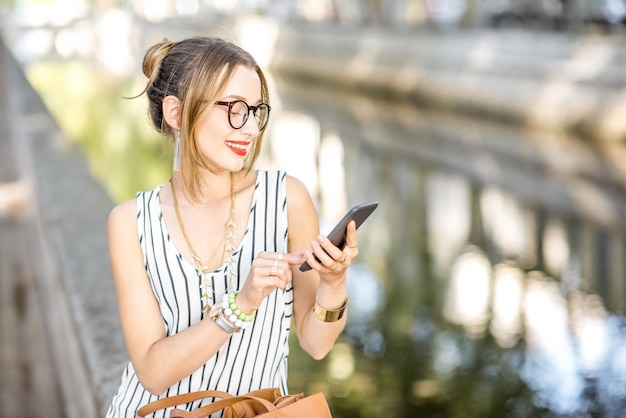 Junge stilvolle Geschäftsfrau, die während des sonnigen Tages mit Telefon und Tasche draußen im schönen Park mit Fluss sitzt