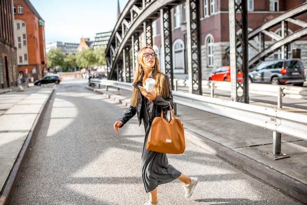 Junge stilvolle Geschäftsfrau in Lederjacke, die die Straße mit Kaffeetasse und Tasche auf der alten Eisenbrücke überquert?