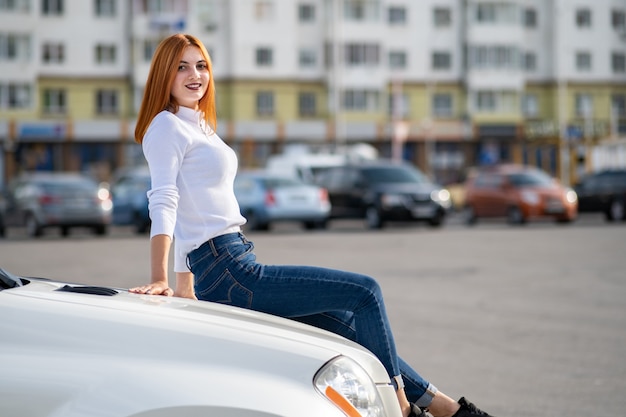 Junge stilvolle rothaarige Frau im weißen Pullover, in den modischen blauen Jeanshosen und in den schwarzen Turnschuhen, die auf einer Motorhaube des neuen teuren Autos auf Stadtstraße sitzen, die warmen Sommertag genießt.