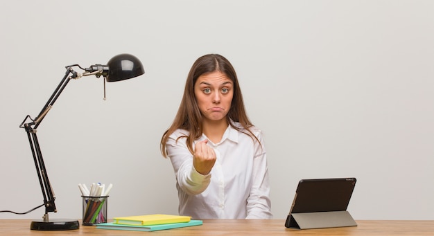 Junge Studentenfrau, die an ihrem Schreibtisch zeigt Faust zum vorderen, verärgerten Ausdruck arbeitet