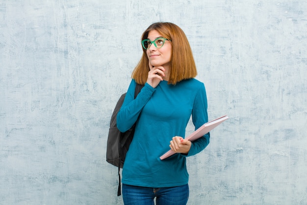 Junge Studentenfrau, die glücklich lächelt und träumt oder zweifelt und zur Seite gegen Schmutzwandwand schaut