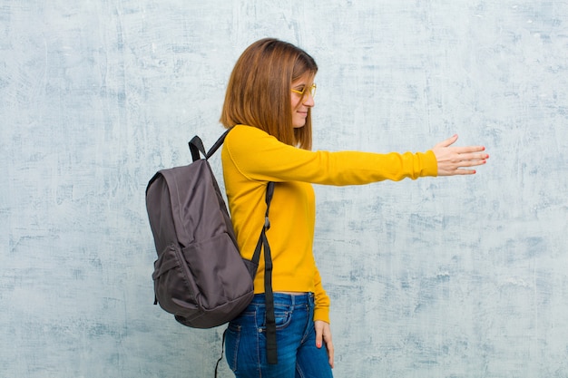 Junge Studentenfrau, die Sie lächelt, grüßt und einen Händedruck anbietet, um ein erfolgreiches Abkommen, Zusammenarbeitskonzept gegen Schmutzwandhintergrund zu schließen