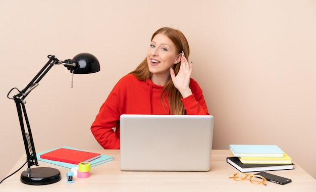 Junge Studentin an einem Arbeitsplatz mit einem Laptop, der etwas hört, indem man Hand auf das Ohr legt