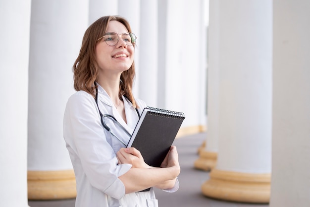 Junge Studentin an einer medizinischen Universität, die im Korridor steht, Porträt einer attraktiven Krankenschwester nahe dem Krankenhaus, glückliche Ärztin mit einem Stethoskop in Uniform