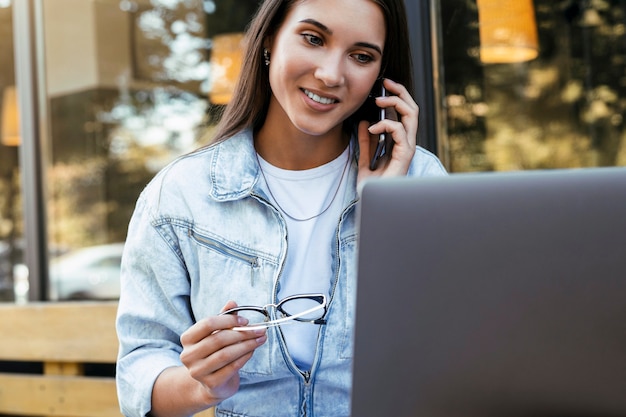 Junge Studentin arbeitet im Café auf einer offenen Terrasse, sitzt vor dem Laptop und spricht am Telefon.