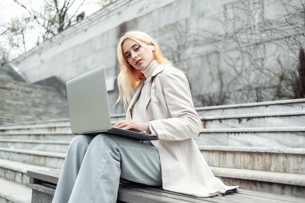 Junge Studentin benutzt einen Laptop, während sie auf einer Bank im Hintergrund der Treppe sitzt