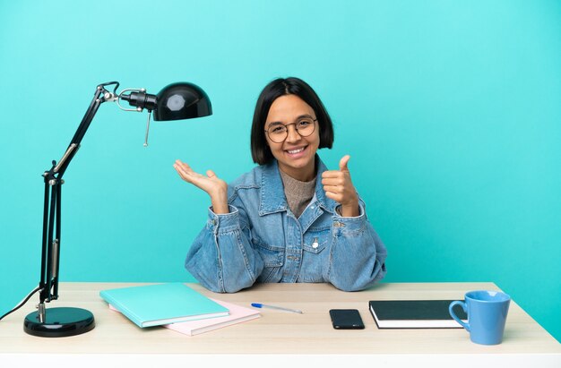 Junge Studentin der gemischten Rasse, die auf einem Tisch studiert, der imaginären Copyspace auf der Handfläche hält, um eine Anzeige und mit Daumen hoch einzufügen