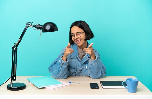 Junge Studentin der gemischten Rasse, die auf einem Tisch studiert, der nach vorne zeigt und lächelt