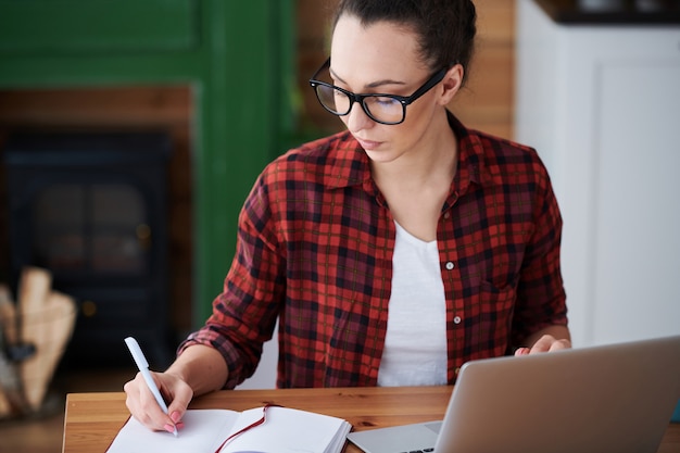 Junge Studentin des Online-Studiengangs macht Notizen, während sie am Tisch vor dem Laptop zu Hause sitzt