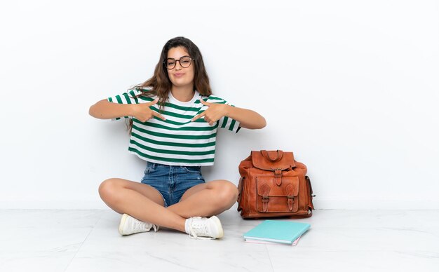 Junge Studentin, die auf dem Boden sitzt, isoliert auf weißem Hintergrund, stolz und selbstzufrieden