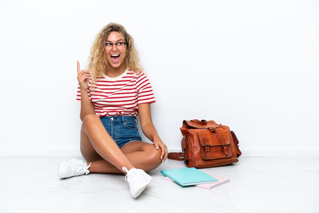 Junge Studentin, die auf dem Boden sitzt und beabsichtigt, die Lösung zu realisieren, während sie einen Finger nach oben hebt