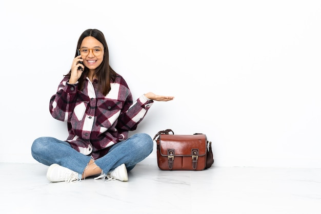 Junge Studentin, die auf dem Boden sitzt und ein Gespräch mit dem Mobiltelefon mit jemandem hält