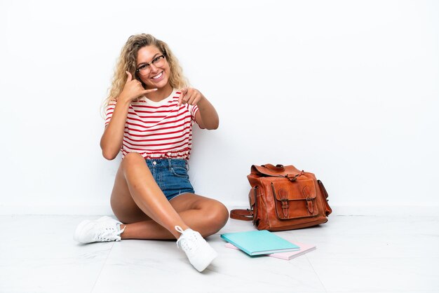 Junge Studentin, die auf dem Boden sitzt und eine Telefongeste macht und nach vorne zeigt