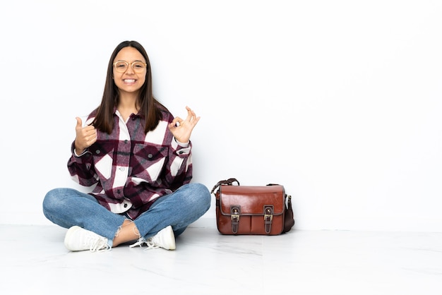 Junge Studentin, die auf dem Boden sitzt und ok Zeichen und Daumen hoch Geste zeigt