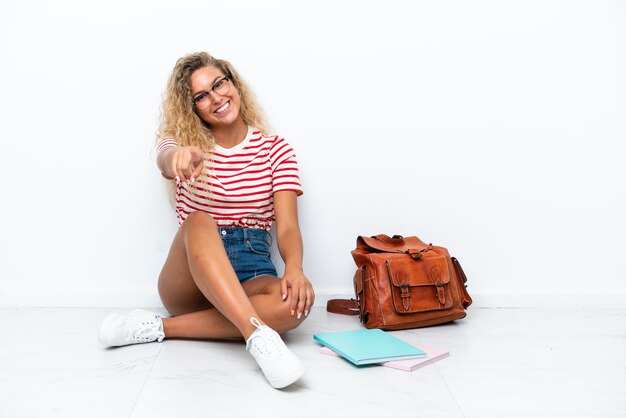 Junge Studentin, die auf dem Boden sitzt, zeigt mit einem selbstbewussten Ausdruck mit dem Finger auf dich