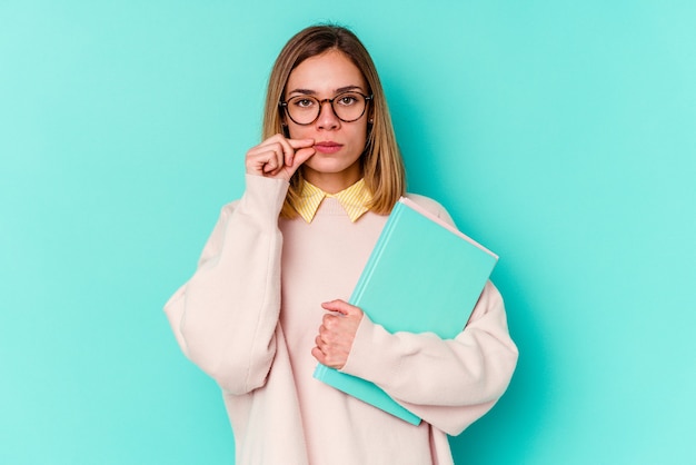 Junge Studentin, die Bücher lokalisiert auf blauer Wand mit Fingern auf Lippen hält, die ein Geheimnis halten.