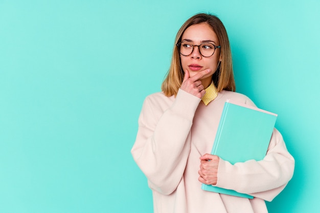 Junge Studentin, die Bücher lokalisiert hält, die seitlich blau mit zweifelhaftem und skeptischem Ausdruck schauen.