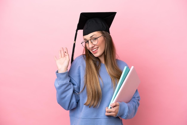 Junge Studentin, die einen Absolventenhut trägt, der auf rosa Hintergrund isoliert ist und mit der Hand mit glücklichem Ausdruck salutiert