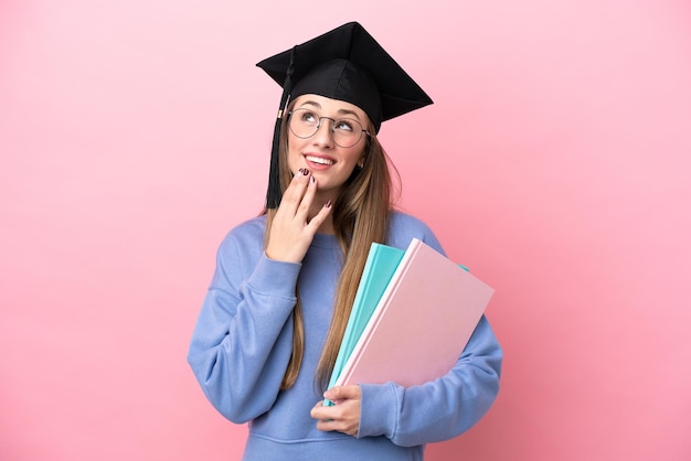 Junge Studentin, die einen Diplomhut trägt, der isoliert auf rosafarbenem Hintergrund aufblickt, während sie lächelt