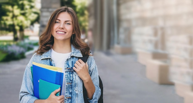 Junge Studentin, die gegen die Universität lächelt, hält Ordner und Notizbücher in den Händen