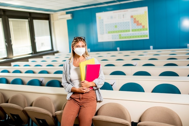 Junge Studentin, die medizinische Gesichtsschutzmaske für Virenschutz trägt, die am Hörsaal steht