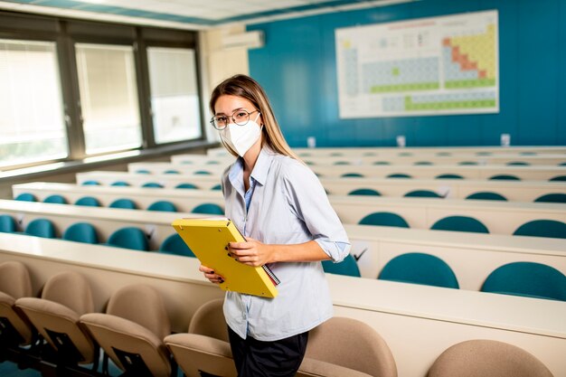 Junge Studentin, die medizinische Gesichtsschutzmaske für Virenschutz trägt, die am Hörsaal steht