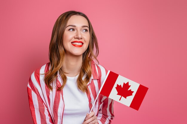 Junge Studentin, die rot gestreiftes Hemd trägt, das lächelt und eine kleine Kanada-Flagge hält und weg lokalisiert über rosa Raum, Kanada-Tagesfeier schaut