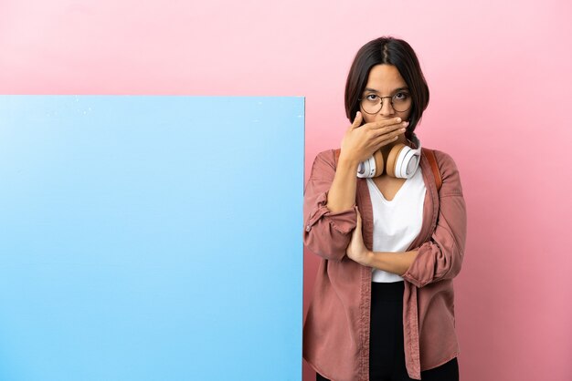 Junge Studentin gemischter Abstammung mit einem großen Banner über isoliertem Hintergrund, der den Mund mit den Händen bedeckt